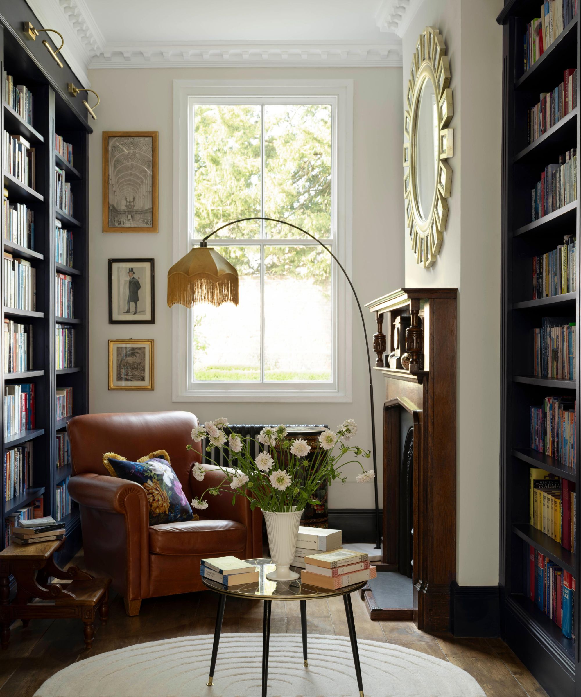 Reading nook with large windows, bookshelves on either side, a leather brown armchair, a vintage floor lamp with a fringed shade, and a round coffee table holding books and a vase of flowers.