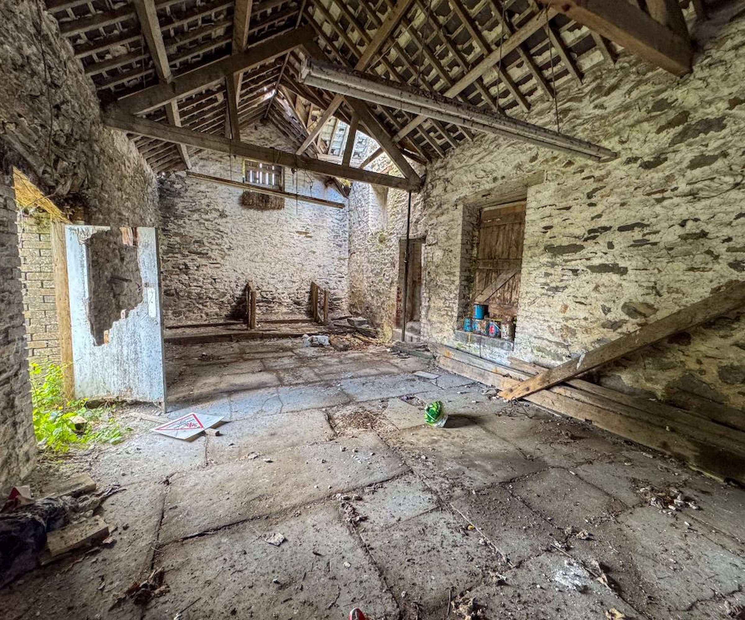 Inside a barn that is up for auction with stone flooring and wooden beams and a broken door and hole in the roof