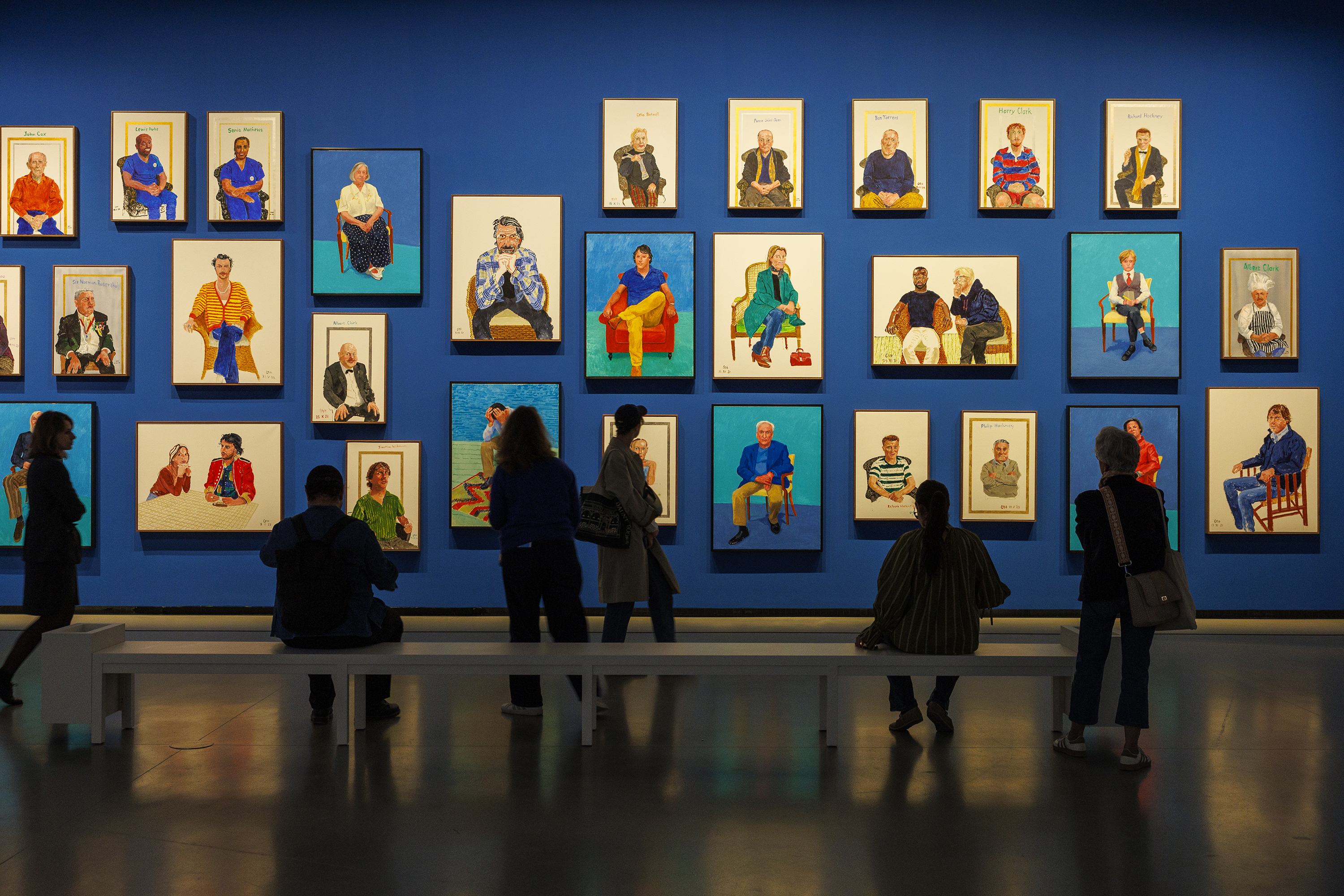 David Hockney's exhibition in Paris — people stand in front of Hockney portraits displayed on a deep blue wall