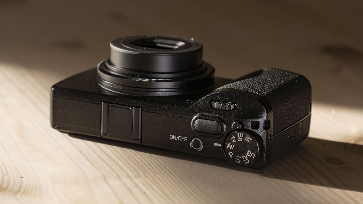 Ricoh GR IV compact camera on a wooden table, facing up, lens active