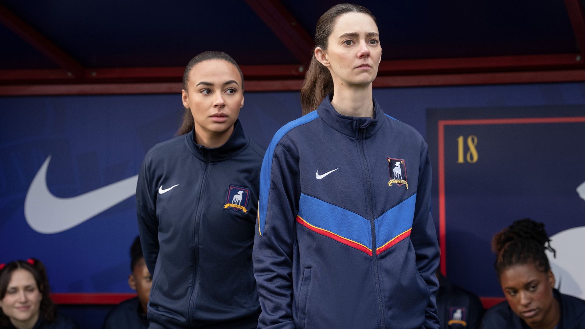 From left to right: Gemma standing right behind Alice on the sidelines during a match on Ted Lasso.