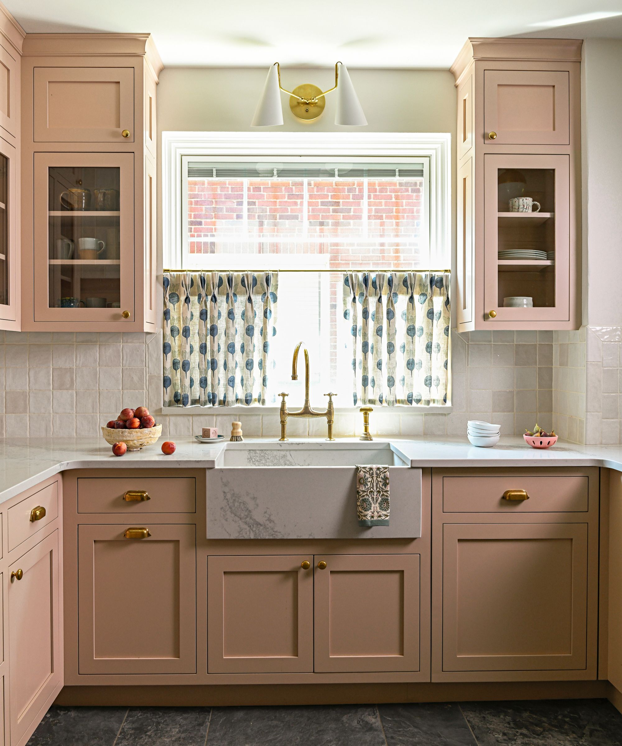 Pale pink kitchen cabinetry with marble countertops and a marble Belfast sink with brass hardware and taps. A window above the sink with patterned cafe curtains.