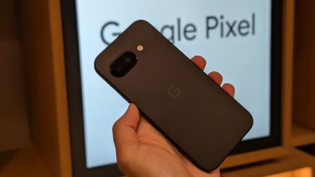 The Obsidian Google Pixel 9a held in hand, backlit by a monitor with the words "Google Pixel" flashing behind it.