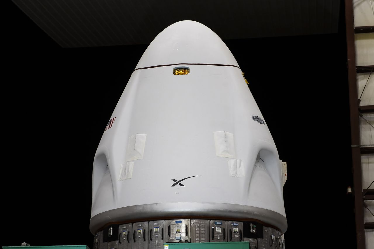 A white conical space capsule stands atop a metal support against a black sky background.