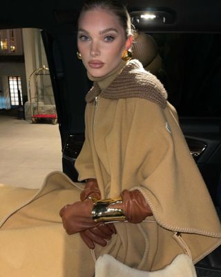 Elsa Hosk in a car wearing a camel-colored cape and skirt with brown gloves and two Elsa Peretti Bone Cuff bracelets by Tiffany & Co.