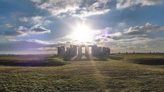 the sun shines through a blue cloudy sky over stonehenge surrounded by rolling grassy fields