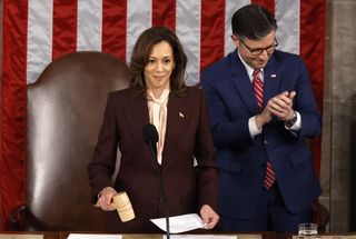 Kamala Harris wearing a maroon suit standing in front of a mic and American flag