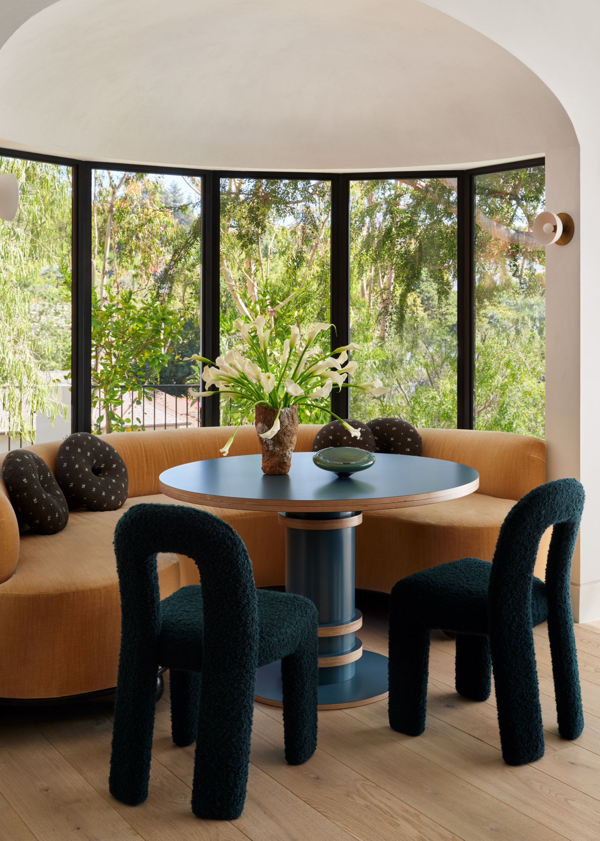 A dining room with curved windows, a wall sconce above a blue round dining table with a ceramic bowl, a vase of flowers, an ochre couch, round patterned windows, and a pair of dark green fuzzy chairs