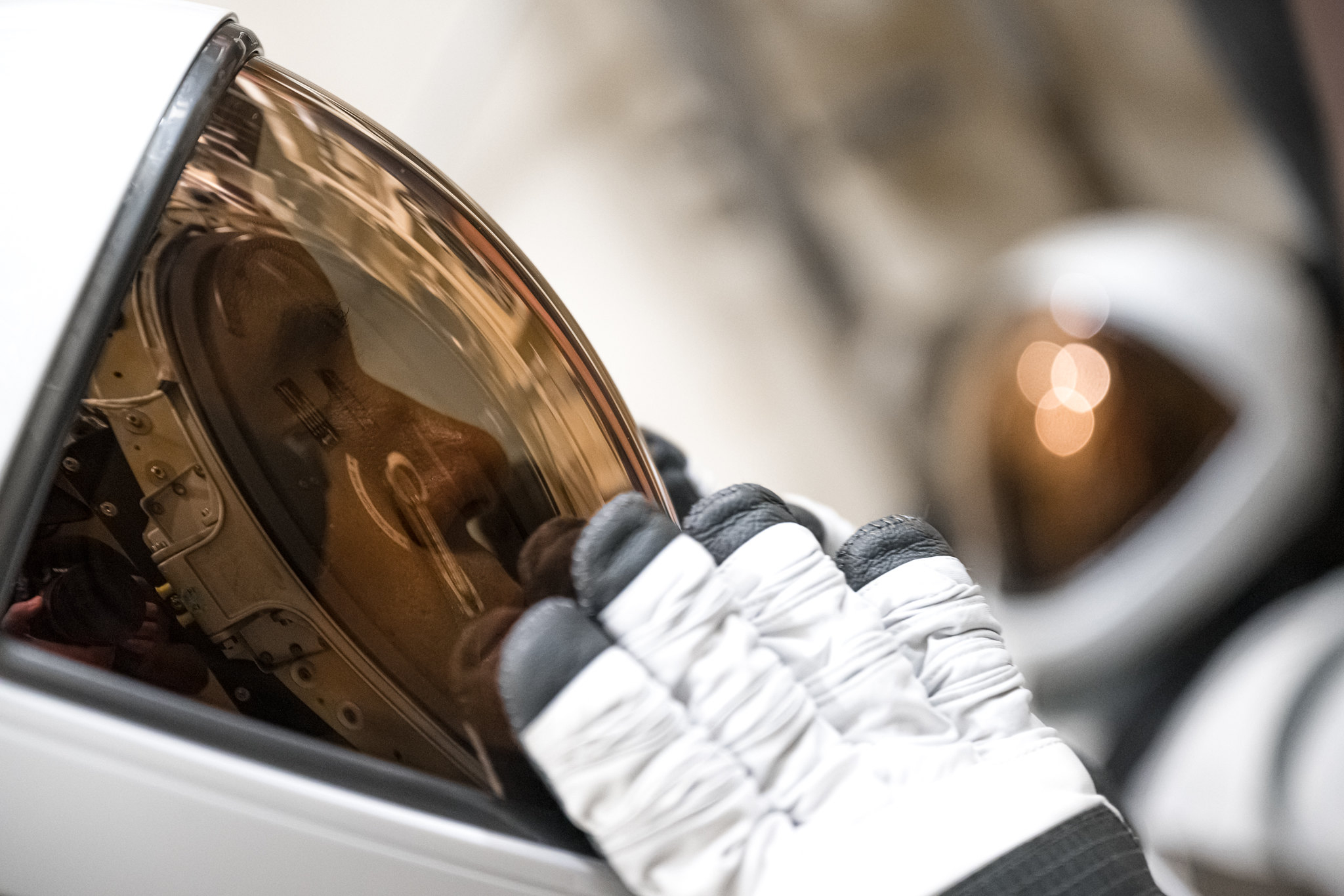 A close-up from the left of a white-shell astronaut helmet that stretches to the center of the image. A copper-shaded visor spans the entire dark interior surface. A pair of black-tipped white gloves is tested on the visor.