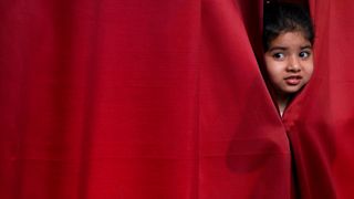 Little girl peeking out from a red stage curtain with a nervous expression