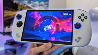 Lenovo Legion Go S gaming handheld in white, running Windows 11, held in front of a blurred gaming room background