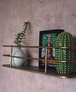 Brass shelf display unit against pink concrete wall with printed wall art, plant and ceramic cactus