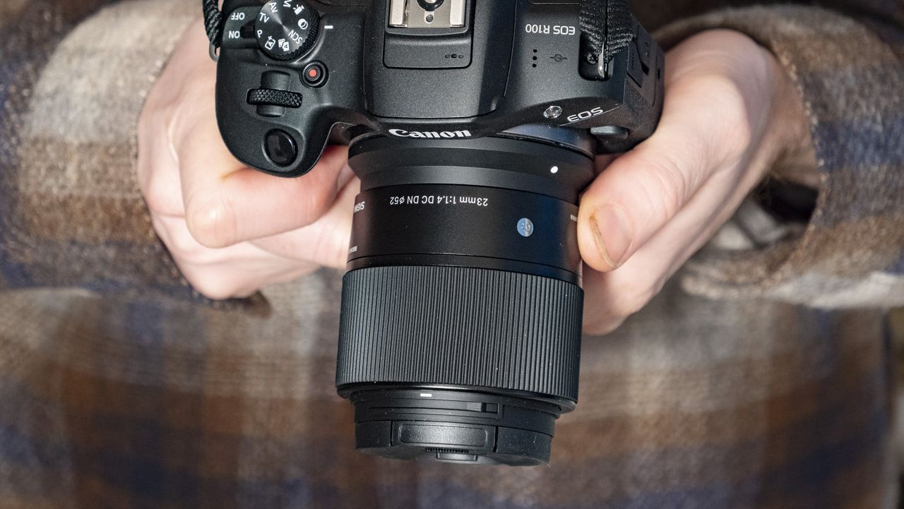 Sigma 23mm F1.4 DC DN Contemporary lens in the hand attached to a Canon EOS R100, checked shirt background