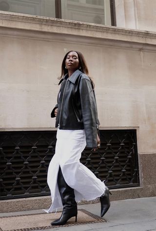 a photo showing the fall jacket trend, cropped leather jackets worn by a woman standing against a tan wall wearing a cropped black leather jacket with a white denim maxi skirt and black knee-high boots