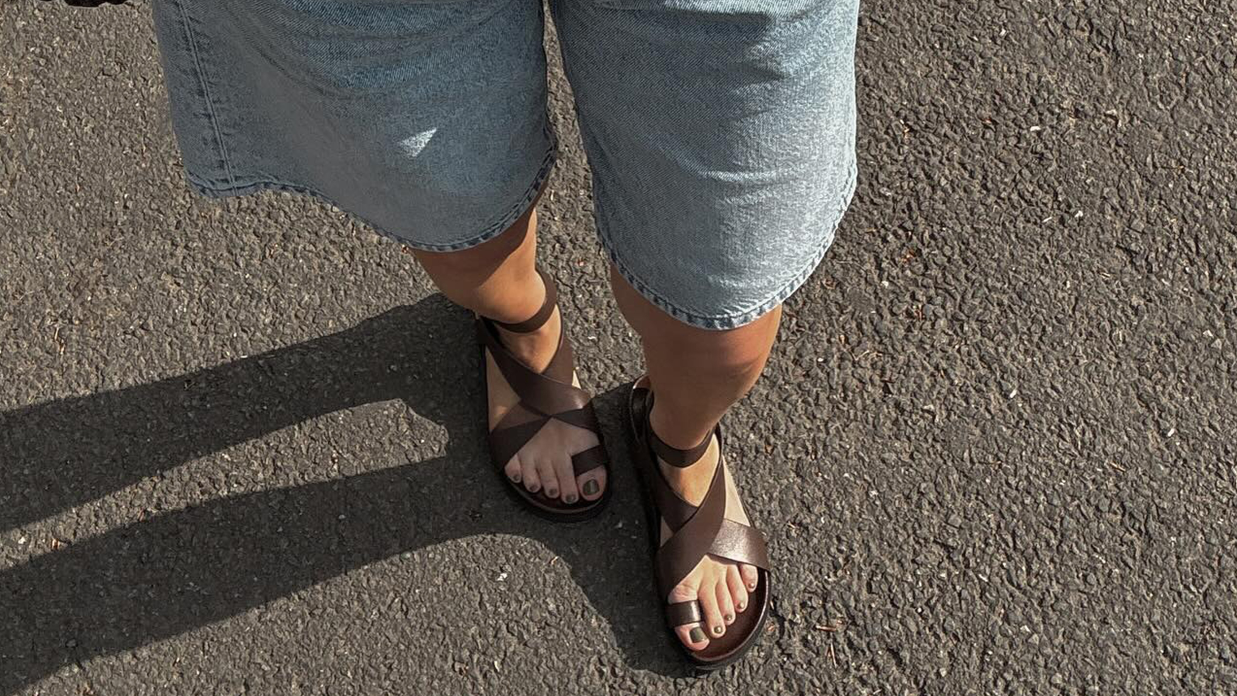 Woman wears A. Emery Jalen leather sandals, Arket long shorts, a black Loewe tank top and a Dragon Diffusion bag while walking.