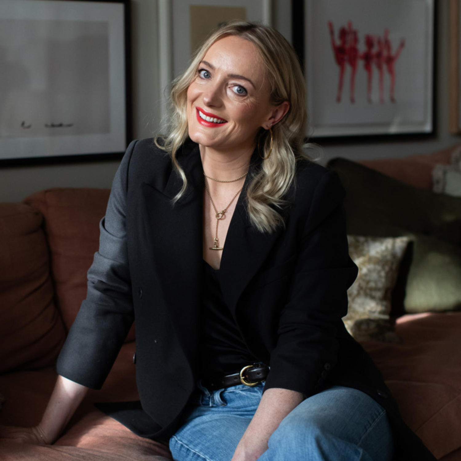 A headshot of a woman with blonde hair and red lips in a black blazer and jeans on a dark brown couch