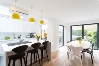A side return extension with an open plan kitchen diner, with all white fittings, yellow hanging lights, and black dining chairs at the island