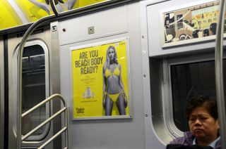 A poster with a woman in a bikini reading 'Are you beach body ready?' in a subway carriage