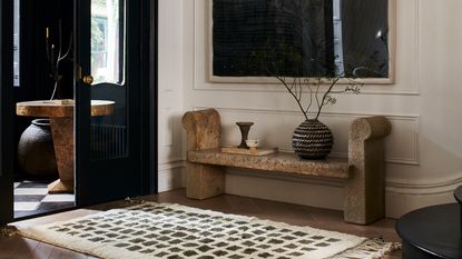 A modern entryway in a Brooklyn townhouse with a bench with decor items on top below a mirror on the wall, a rug and a set of black doors leading to the porch with a round table and vase in it