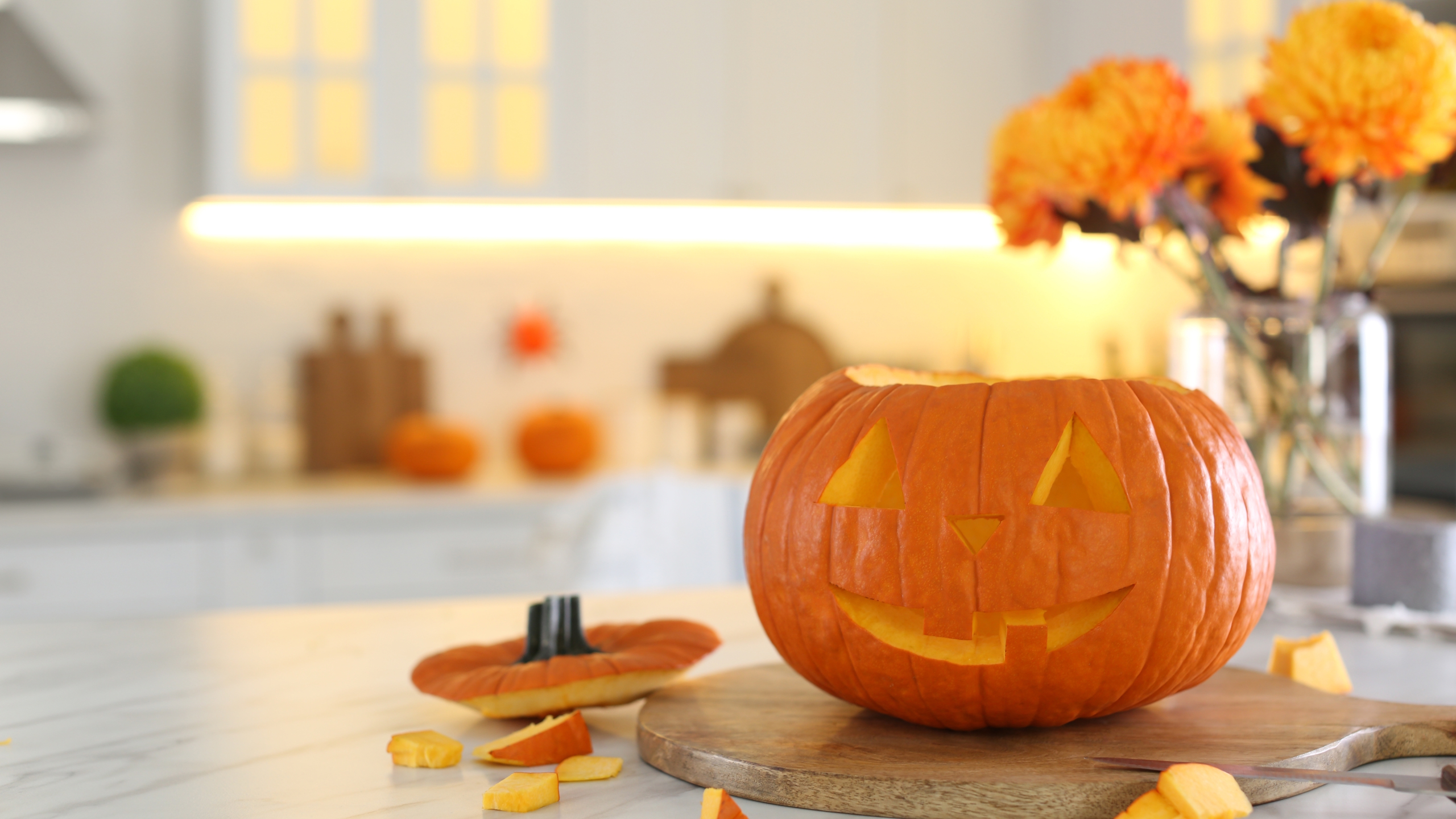 A carved pumpkin sits on a wooden board on a white marble kitchen top, with orange flowers in the background