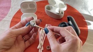 Hands holding a set of earbuds and a Q-tip to clean them, with other earbuds and cases in the background