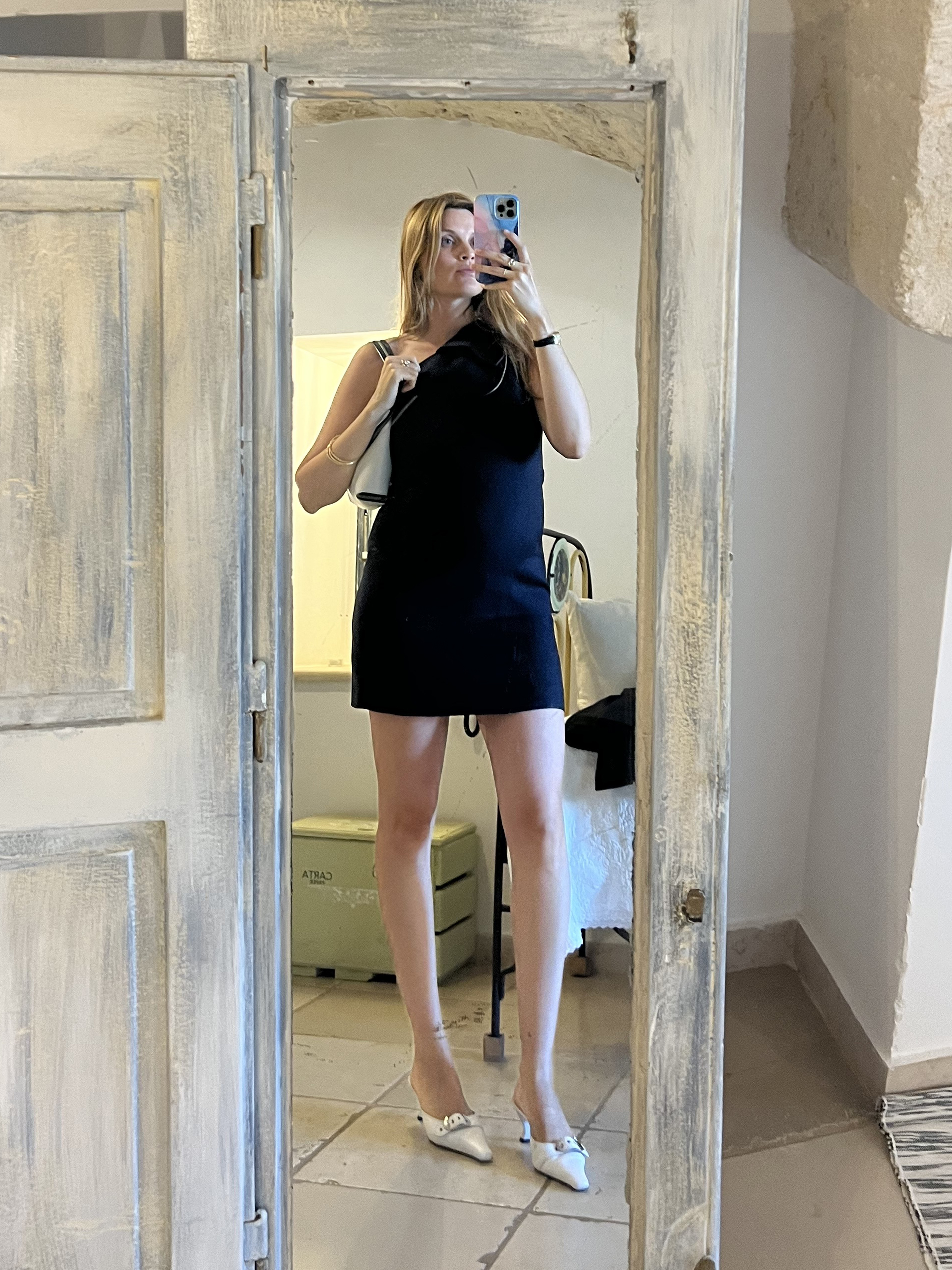 French editor and fashion creative Eugenie Trochu poses for a mirror selfie wearing a black Versace mini dress, a white shoulder bag, and white By Far buckled mule kitten heels