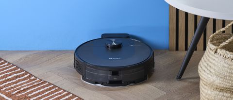 a rechargeable robot vacuum and mop in black by ultenic is photographed on a hardwood floor with a blue wall and a burnt orange terracotta and white striped rug