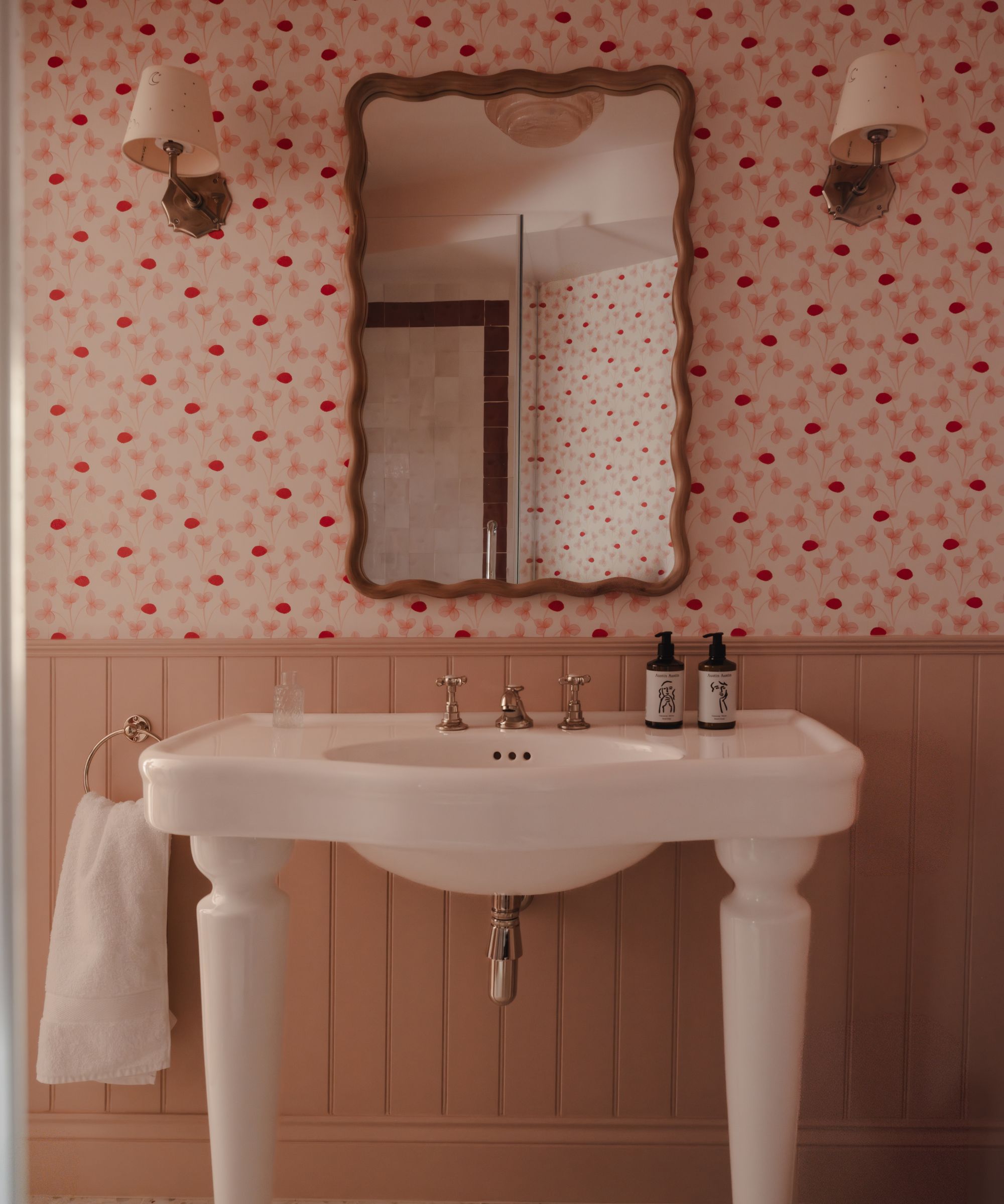 a pink bathroom with half height paneling, floral wallpaper and a traditional ceramic vanity