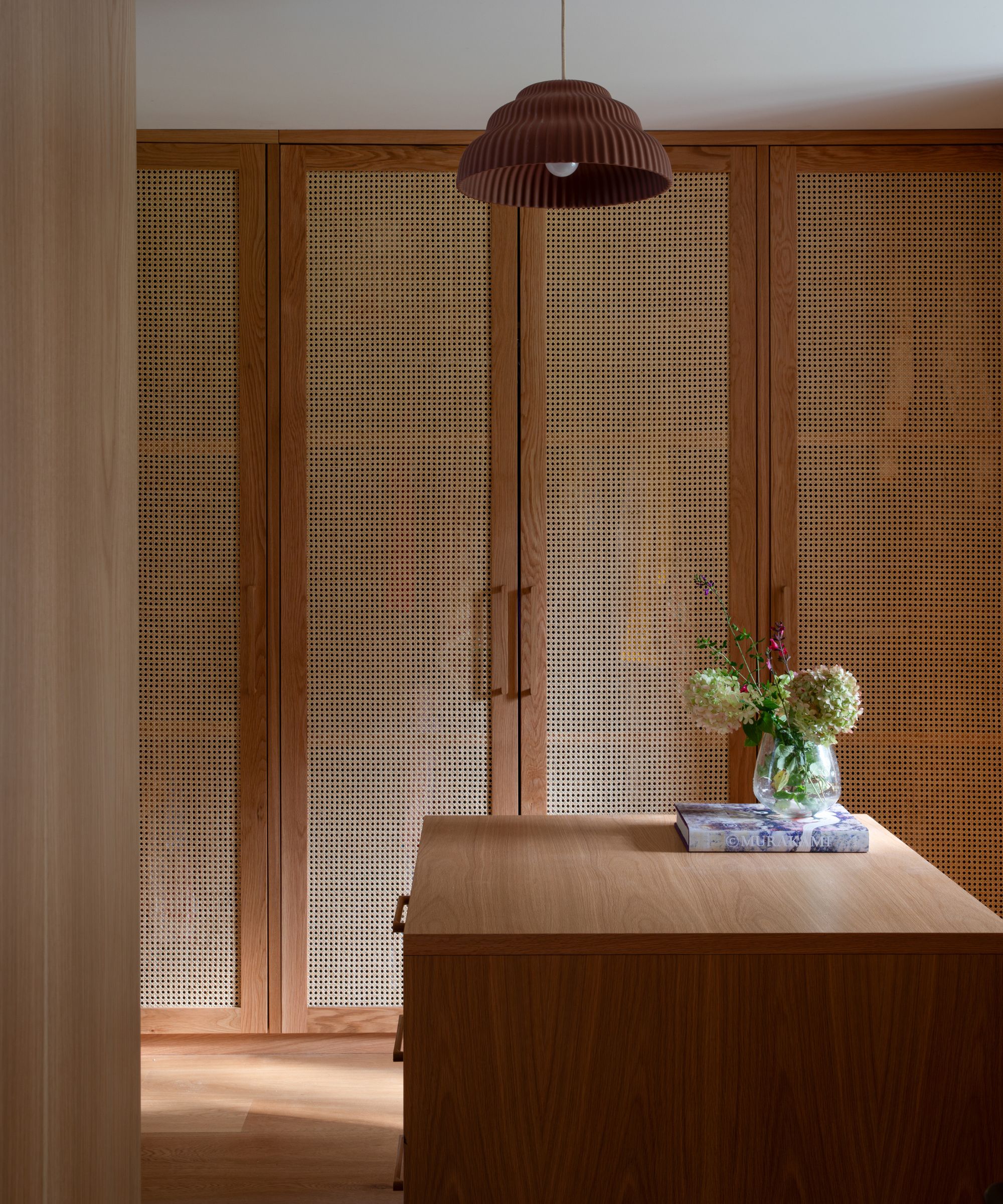 Built-in wall of wooden closets with rattan door fronts. A wooden island in the centre of he room with a small vase of flowers, and a rattan pendant light hanging overhead.