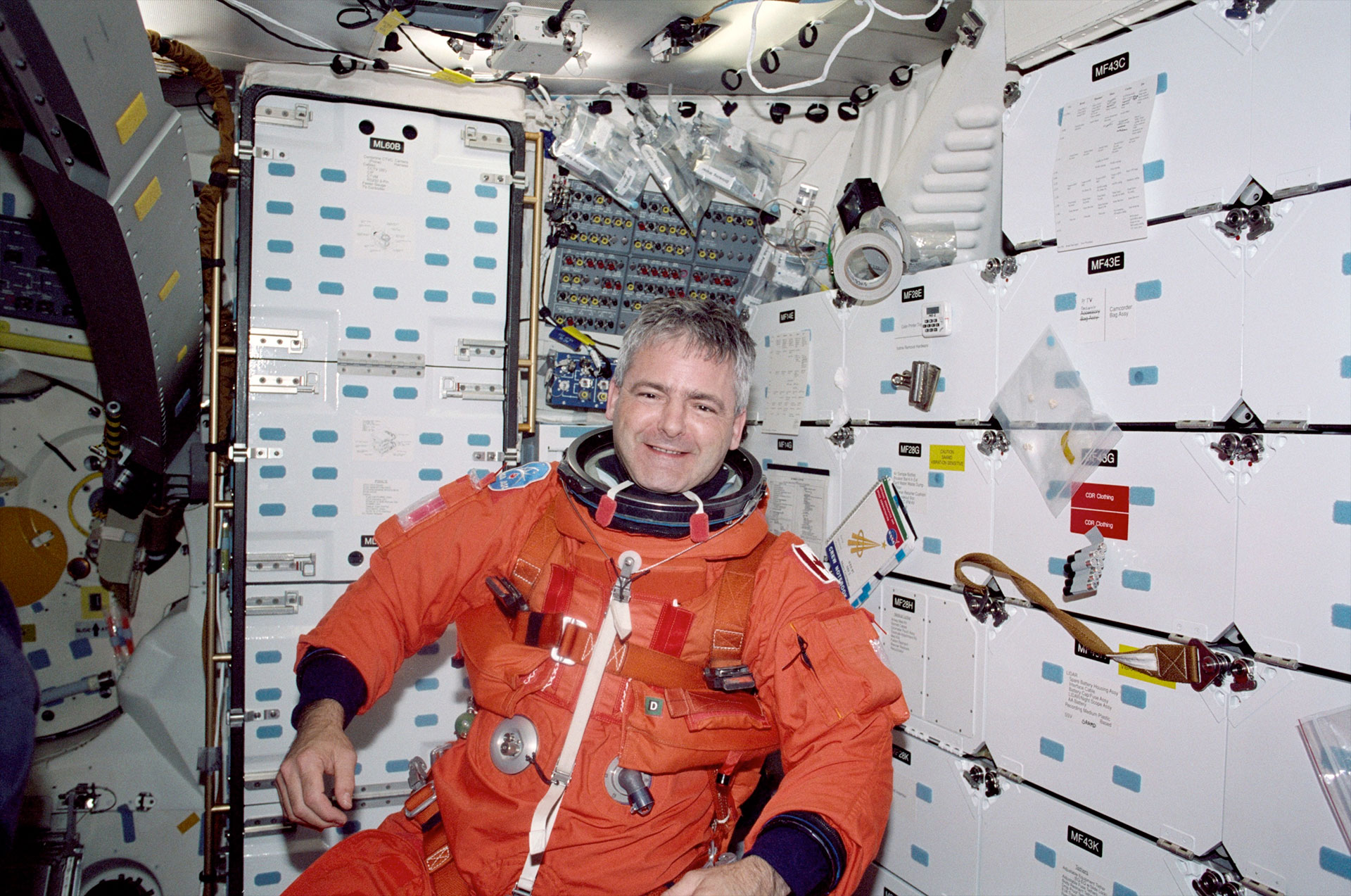 a man in a bright orange astronaut pressure suit floats inside a spacecraft crew cabin.
