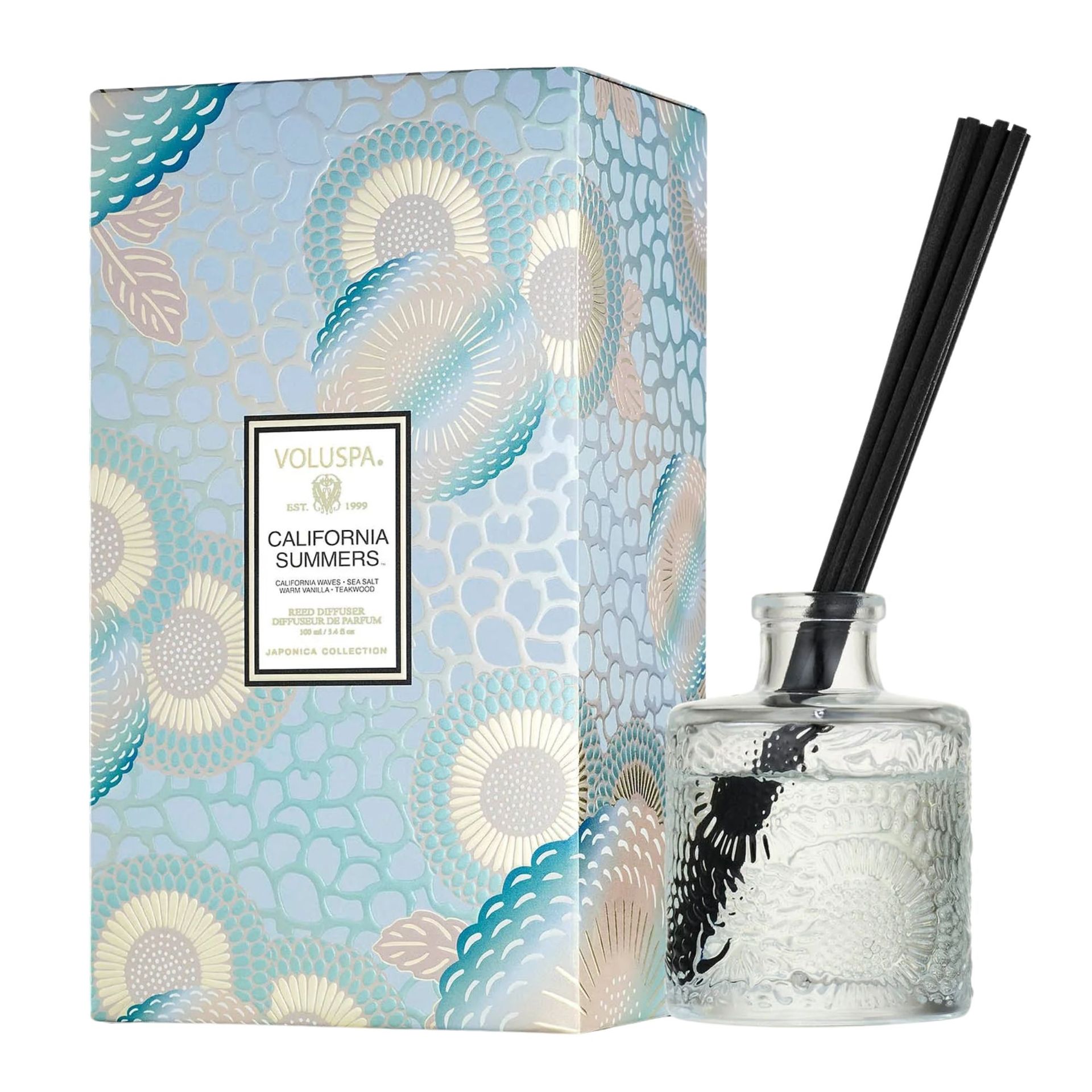 A small glass reed diffuser with black reeds next to a blue patterned packaging box