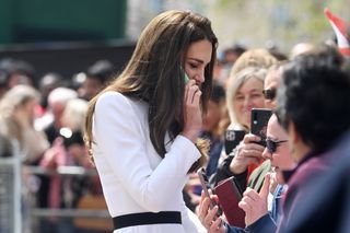 Kate Middleton wearing a white dress with a black belt and talking on a member of the public's cell phone during the coronation as people take her photo