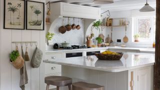 A charming country-style kitchen featuring a white marble-topped island with wooden stools. The space includes open shelving, brass hanging rails, and a backsplash with a herringbone tile design.