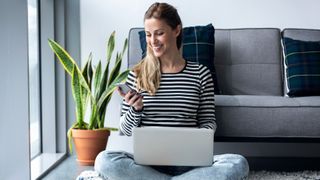  woman using her mobile phone while working with laptop sitting on the floor at home.