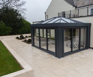 square black framed conservatory attached to large cream house with cream stone paved exterior area