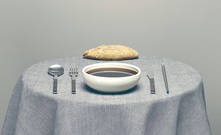 A round table with a grey table cloth. A white porcelain bowl, cutlery and a bread roll.