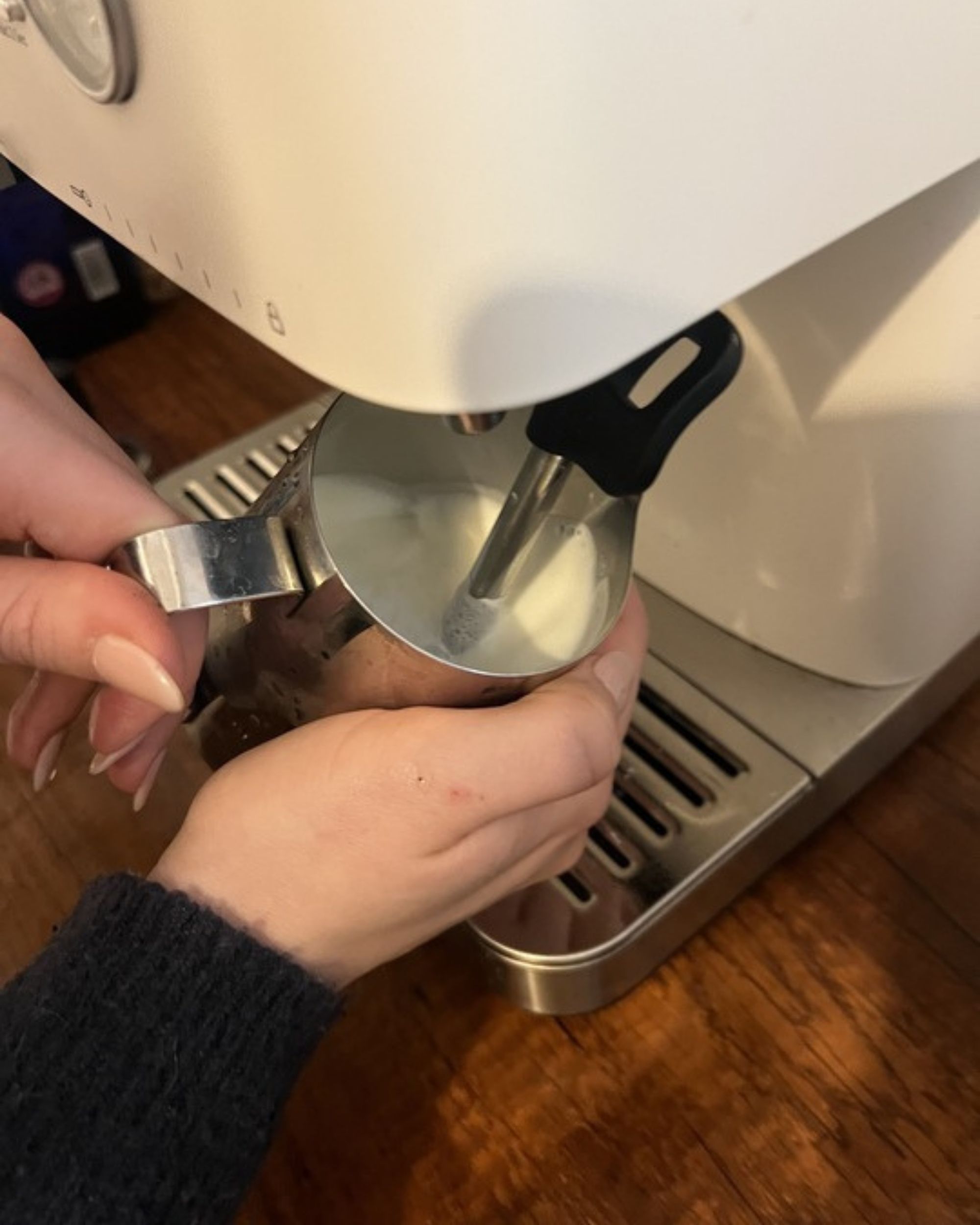 Image of a hand holding a milk jug under a steaming wand attached to a white espresso machine.