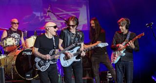 G3 Tour 2024 [L-R]: Joe Satriani, Steve Vai, and Eric Johnson jam onstage as the three instrumental powerhouses united for a triumphant tour. The legendary Kenny Aronoff plays drums in the background.