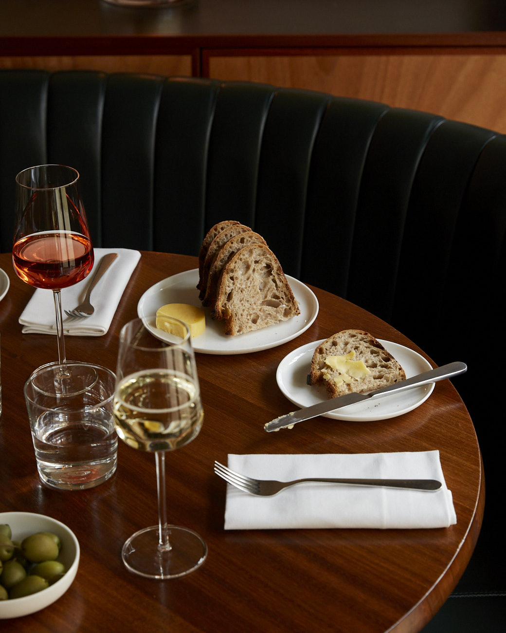 A sleek round wooden table prepared for diners shows two plates of bread and butter, wine glasses, and bowls filled with olives and a leathery blue banquette.