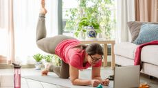 A woman practices Pilates at home in her living room on a mat. She is balancing, close to the ground, on her forearms and her bent left knee. Her right leg is bent at 90° and elevated behind her. In the room we see a wtaer bottle, a couch, a plant and a coffee table. A laptop is open in front of her.
