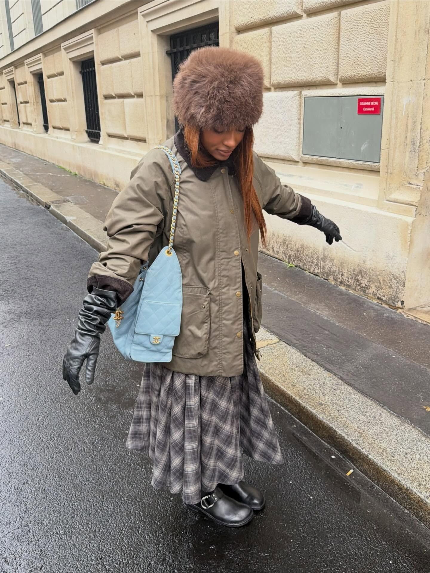 MV Tiangue wearing a gray plaid skirt with a brown chore jacket, fluffy hat, a blue Chanel handbag, and black clogs.