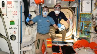 two men in short sleeve shirts and kahki slacks wear protective glasses, masks and gloves to protect against floating debris before entering a cargo ship berthed with a space station.