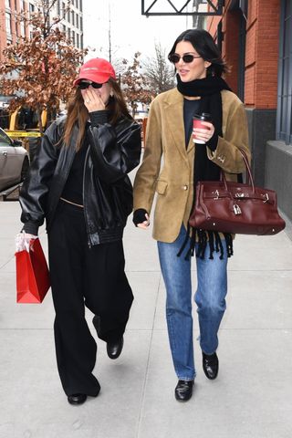 Hailey Bieber wears a red hat, black bomber jacket, black top, and black pants. Kendall Jenner wears a black scarf, black top, brown suede blazer, burgundy Hermes bag, jeans, and loafers.