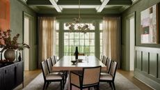 green dining room with coffered ceiling, large dining table with chairs, patterned rug, black console with orange artwork hanging above, and curtains partially covering wall of windows