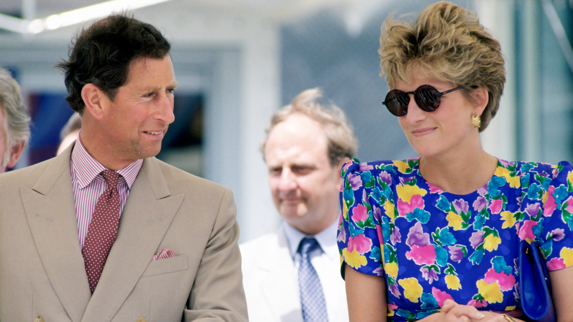 King Charles looks at Princess Diana who is wearing a blue floral dress featuring pink, yellow, and purple flowers with round sunglasses