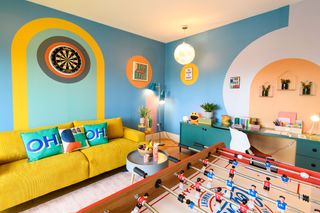 Brightly colored room with blue, yellow and orange accents, a table football, yellow couch and graphic cushions