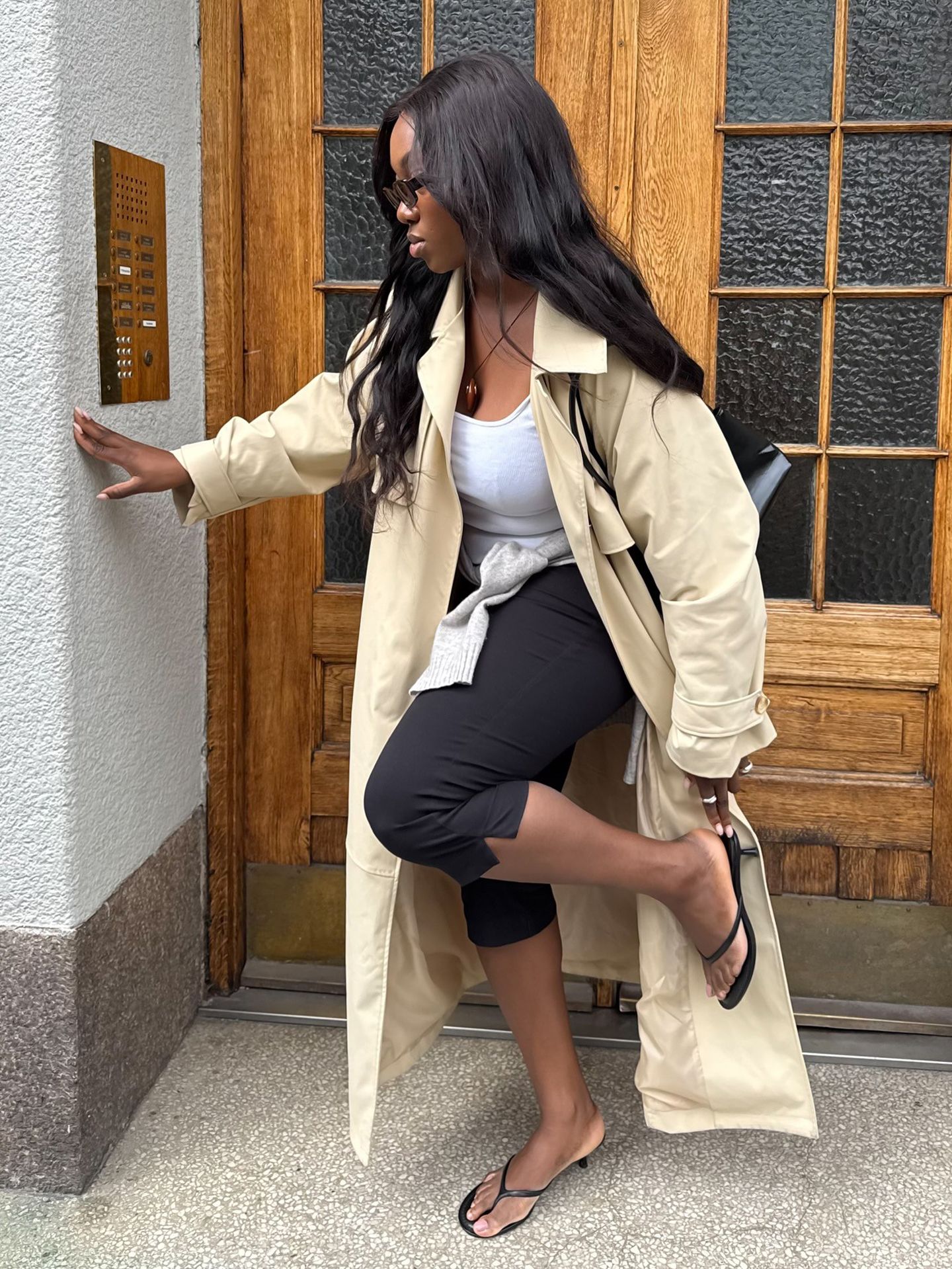 style influencer Nnenna Echem poses wearing a trench coat, white top, cord necklace, black capri pants, and black flip-flop kitten heels