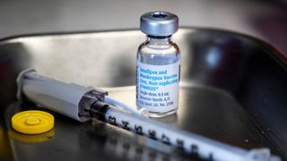 photo of a labeled vial of the jynneos mpox and smallpox vaccine on a metal tray next to a syringe with a protective cap over its tip