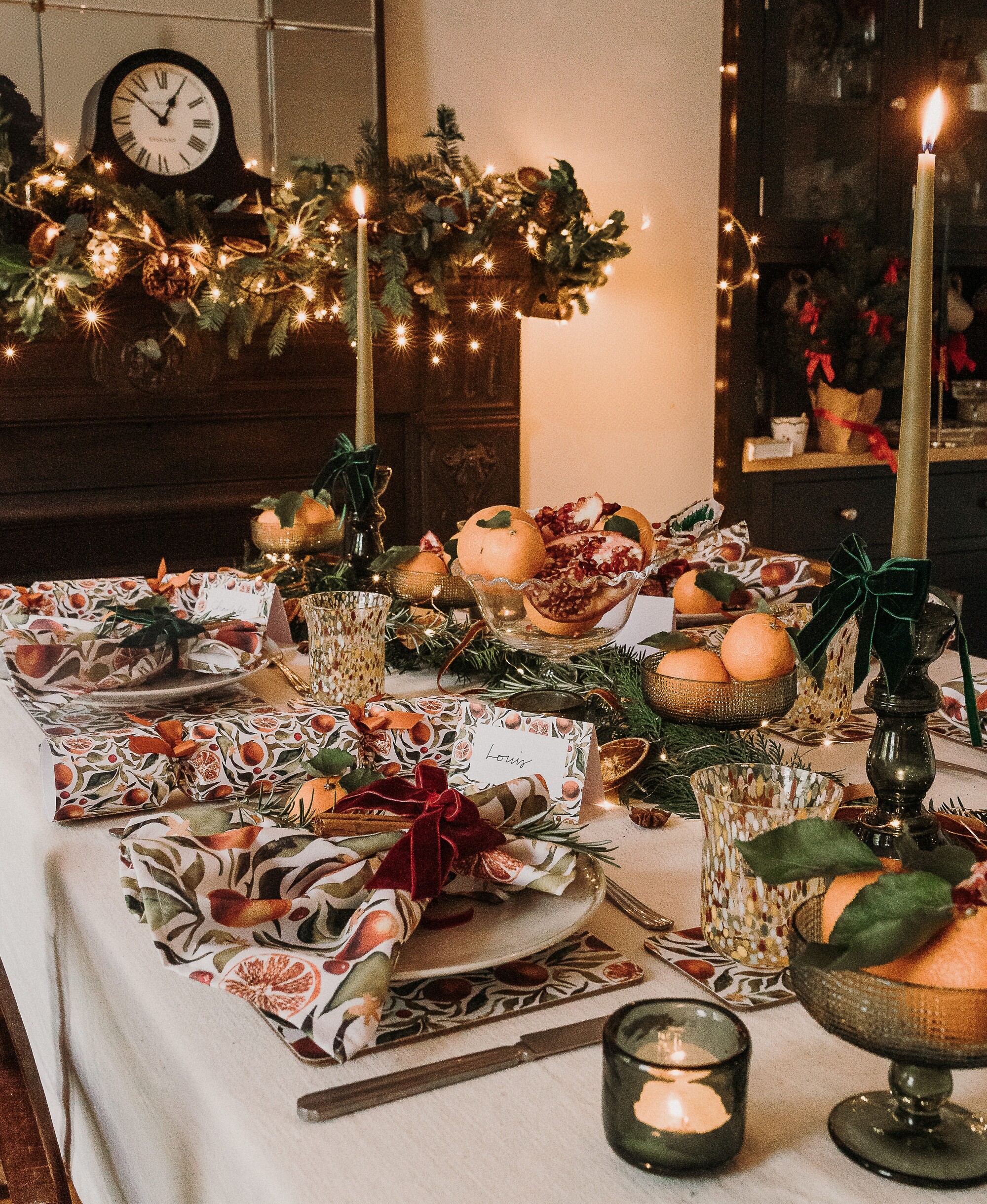 christmas table setting with patterned orange-motif napkins, green glassware, tall candlesticks, next to an ornate fireplace with garland