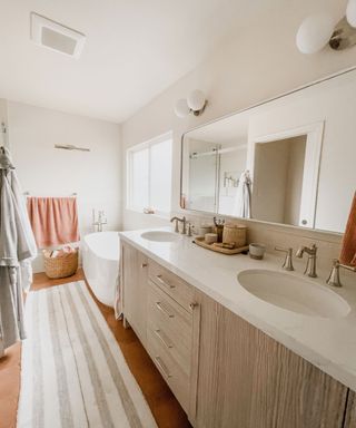 A white and brown bathroom with pink towels, a runner rug, bath, and dual sink